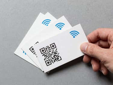 Overhead flat-lay of three physical business cards fanned on a matte gray surface, one card face-up showing a QR code grid and NFC wave icon printed in the corner, a thumb entering frame from the right about to tap, clean daylight from above, sharp detail on the card stock texture