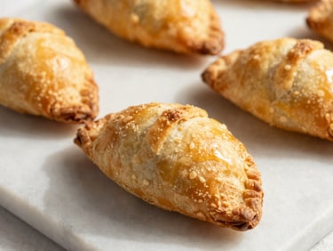 Golden brown puff pastries and hand-pies arranged on a clean marble slab, warm Miami daylight casting soft shadows, flaky texture close-up.