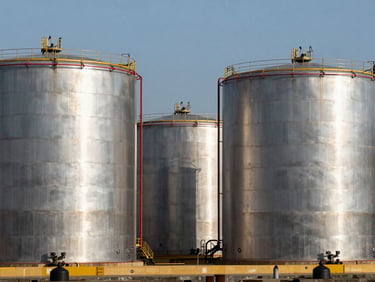 Bulk liquid storage tanks at a maritime port, reflecting sharp daylight off steel surfaces.