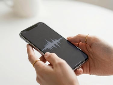 Close-up macro shot of elegant hands with delicate gold rings holding a glowing smartphone displaying an audio waveform, bright natural daylight, soft focus.