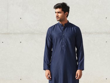 A young South Asian man in a modern navy blue panjabi with minimal embroidery, standing against a bright, minimalist concrete wall, soft daylight.