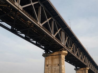 Dramatic low-angle view of massive steel bridge trusses meeting concrete piers under cool overcast daylight, emphasizing engineering precision.