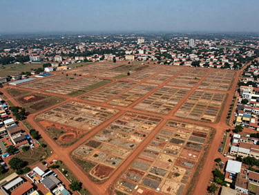Wide aerial drone photograph of a planned urban district in Cameroon under development — grid of plotted land parcels visible from above, red-earth roads cutting geometric patterns, sparse emerging construction, blue sky above