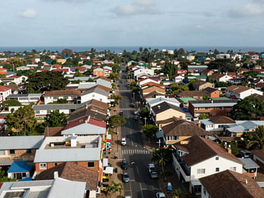 Wide aerial of Osu commercial and residential mix — Oxford Street corridor visible with rooftop signage below, Gulf of Guinea coastline on the far horizon, natural daylight