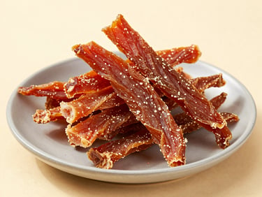 Close-up studio shot of a pile of dehydrated chicken jerky strips on a soft grey ceramic plate, warm cream background, high-key daylight, texture and grain clearly visible