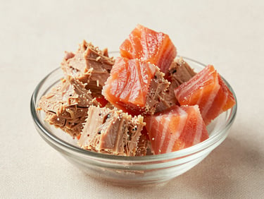 Close-up studio shot of tuna crunch bites and salmon treat cubes arranged in a small glass dish on a light cream linen surface, high-key studio daylight, clear texture detail