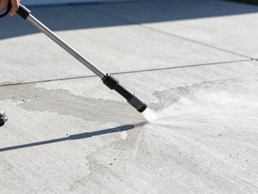 Close-up of a pressure washing wand cleaning a concrete driveway, half clean and half dirty, bright midday sun, sharp contrast, low angle.