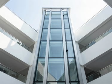 Looking directly up a modern glass elevator shaft inside a bright, sunlit commercial atrium, minimal architecture