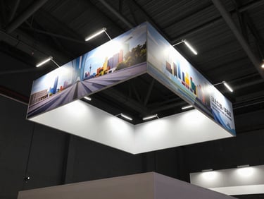 Wide environmental shot of a fully built commercial stand inside a trade fair hall, double-height structure with illuminated graphic panels, angled architectural framing from below showing scale and fabrication craft, deep charcoal hall background, practical booth lighting, no people, no text
