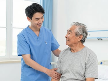 Caring male patient care assistant assisting an elderly patient in a bright hospital room, both smiling. Bright natural daylight, professional environmental portrait, sharp focus.