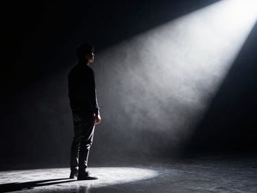 Dramatic silhouette of a speaker standing in the wings of a stage, looking out toward a bright spotlight, deep shadows, high-contrast