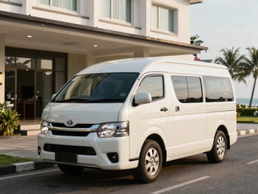 A white Toyota Hiace passenger van parked in front of a modern hotel lobby in Kupang, clean and polished, eye-level shot