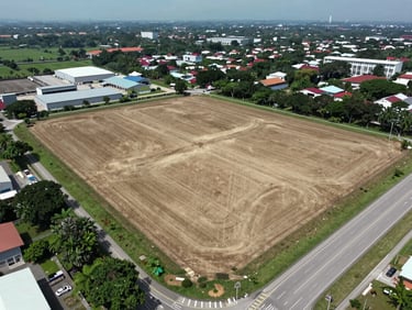 Wide aerial drone shot of undeveloped land parcel adjacent to a main road in Johor Bahru, visible lot perimeter, cleared ground, industrial surroundings, overcast natural daylight, no people
