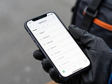Tight crop of an Android phone screen showing a field service scheduling UI, technician's gloved hand gripping the device, overcast outdoor light, tool bag visible at the edge of frame