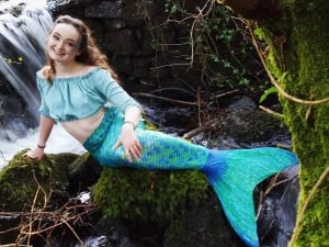 Mermaid Echo wearing a blue tail. She is sitting on a rock.