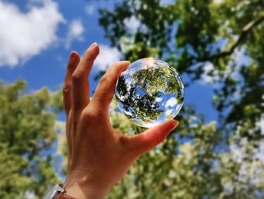 A hand holds a small glass globe reflecting trees and sky, symbolizing the beauty of creation.