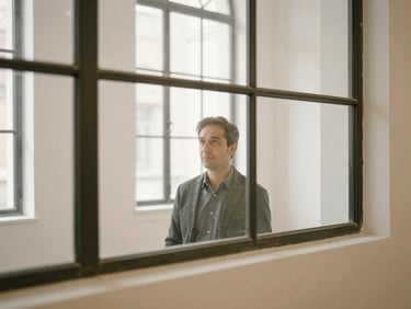 A candid executive portrait of a professional looking through large steel-framed windows in a quiet gallery space.