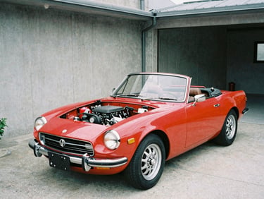 Three-quarter view of a classic red Japanese roadster with soft top down, parked in a tidy concrete garage, engine bay exposed, natural light