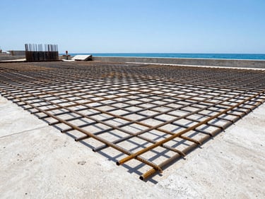 Low-angle heroic shot of a reinforced steel rebar grid on a coastal construction site under bright midday sun, ocean visible in the background.