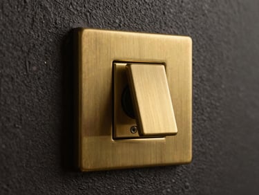 Macro close-up of a premium solid brass light switch integrated perfectly flush into a dark textured plaster wall, warm golden light grazing the metal