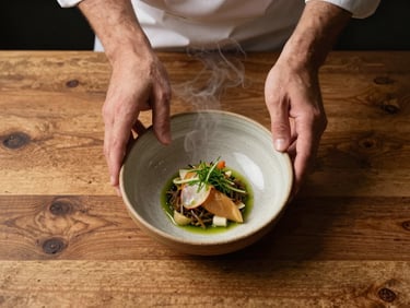 Close overhead flat-lay of a chef's hands plating a dish on a worn wooden counter, warm available interior light, rustic ceramic bowl, visible steam, honest and unposed