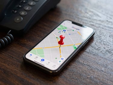 A close-up macro shot of a mobile phone screen displaying a Google Maps interface with a red pin, resting on a dark, unvarnished wooden desk next to a silent office phone, natural daylight.