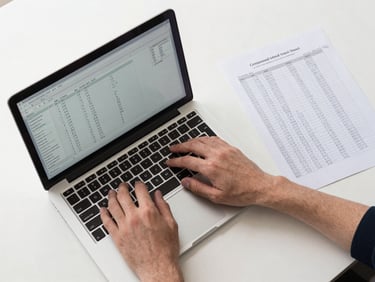 Overhead flat-lay of a desk: hands typing on a laptop keyboard displaying a financial calculator interface with formula fields, a printed spreadsheet with handwritten compound interest calculations visible to the right, soft neutral studio lighting, clean white surface, no shadows