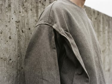 A close-up of a heavyweight cotton garment, architectural drape, dropped shoulder detail, against a brutalist concrete wall, low-contrast overcast daylight, medium-close crop.