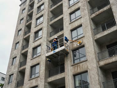 Wide environmental shot of a multi-unit residential building exterior mid-cleaning, worker on a platform scrubbing the facade, water running down the concrete surface, overcast daylight, urban context visible