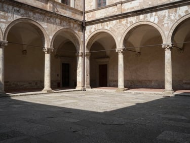 Empty stone courtyard of a historic Italian cultural institution at midday — arched loggia casting long shadows, a closed wooden door, no people, architectural texture showing age and continuity