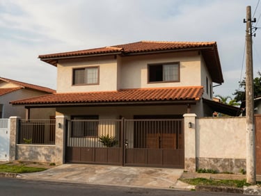 Exterior of a two-story house in Navegantes at mid-morning, terracotta roof tiles, open gate, concrete driveway, overcast natural light, quiet residential street