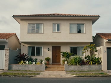 Front view of a two-storey house in Penha, Santa Catarina, photographed from the entry gate, neutral overcast morning light, small garden, tiled façade, quiet residential street visible in foreground