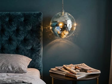 Styled bedroom corner with a disco-ball hanging above a tufted velvet headboard in deep teal, warm golden lamp light, vintage fashion magazines fanned on a side table, rich layered textiles