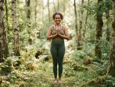 A warm, editorial portrait of a diverse yoga instructor with a kind smile, mid-shot, in a sun-dappled forest clearing, soft diffused light, earthy moss green and birch tones, shallow depth of field.