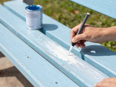 A close-up of a hand painting a weathered wooden park bench with fresh white paint, bright Florida morning light, paint can nearby, 35mm