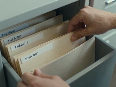 Close-up of hands sorting archival institutional records in a filing system, document folders labeled with academic years, cool available daylight, overhead angle, no faces visible