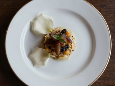 Close overhead view of an elegantly plated wedding dinner course on white porcelain, gold-rimmed charger plate, single white rose petal beside the arrangement, soft studio light