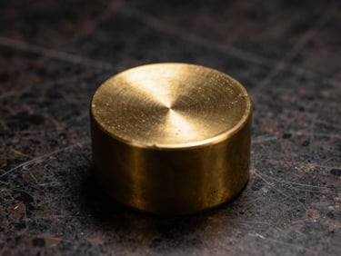 Close-up study of a collectible object — a solid brass paperweight on dark marble, single overhead studio light, sharp specular highlight, surrounding deep shadow, no color cast, monochromatic