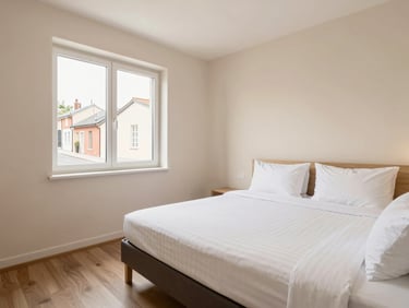 Bright, clean standard double room with a made bed, warm lighting, and a window overlooking a quiet street, wide-angle.