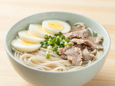 Close-up of a steaming bowl of noodle broth with fresh scallions, sliced soft-boiled egg, and tender pork, bright natural daylight