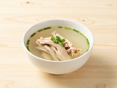 A steaming white bowl of clear ginger chicken broth with scallions and pulled chicken on a light birch wood table