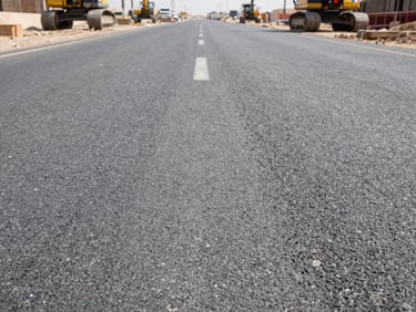 Close-up of a concrete road surface under construction in Saudi Arabia, overcast daylight, heavy machinery visible at the far edge, strong texture of aggregate and fresh asphalt, no people in foreground