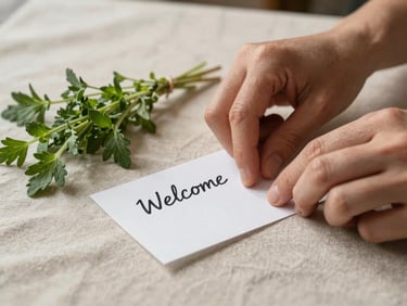 Close-up of hands placing a handwritten welcome note beside a small bunch of fresh herbs on a linen tablecloth, warm north-facing daylight, simple and real