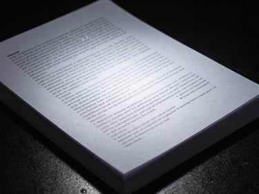 Close-up of dense printed legal document pages stacked at an angle under a single overhead spotlight, dark background, cool directional light, no readable text