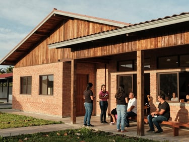 Documentary shot of a modern community center building in Colombia, brick and wood architecture, warm afternoon sun, local residents talking outside.