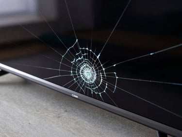 Extreme close-up overhead shot of a cracked LED TV panel, spider-web fracture lines visible under bright studio strobe, black screen, workbench surface below