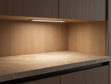 Detail shot of integrated under-cabinet task lighting illuminating a stone worktop below, warm directional light, wood-grain cabinet interior visible in background, no people, tight environmental framing