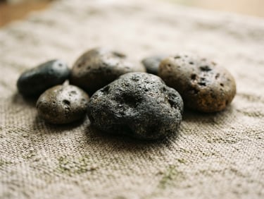 Close-up of warm basalt stones resting on a textured linen surface, soft diffused natural daylight, shallow depth of field, 35mm, warm editorial photography, muted moss green and warm stone tones