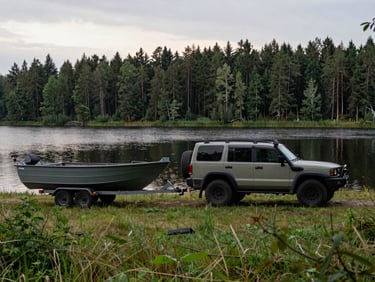 Wide environmental shot of a robust SUV towing a fishing boat on a trailer, parked by a calm Lithuanian lake at dawn, overcast soft morning light, deep forest green and natural tones, 35mm photography.