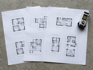 Overhead wide shot of technical project drawings and specification sheets laid flat on a concrete surface with a measuring instrument beside them, overcast natural light, no people visible